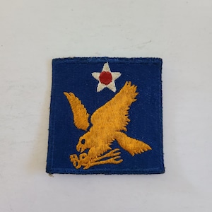 May include: A square, embroidered patch featuring a golden eagle with outstretched wings, clutching a bundle. The eagle is set against a blue background, with a white star and a red center at the top. The patch has a stitched border.