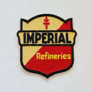 May include: A yellow and red embroidered patch with a black outline. The patch features a shield shape with a cross at the top and the text "IMPERIAL Refineries".