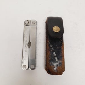 May include: A silver multi-tool with the text "LEATHERMAN TOOL" and a dark brown leather sheath with the text "LEATHERMAN". The multi-tool is open, revealing various tools. The sheath has a gold-colored button.