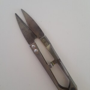 May include: A pair of small, silver-colored thread snips with pointed blades. The snips have a spring-loaded handle and are designed for cutting threads or small cords. The blades are slightly rusted, indicating age or use.