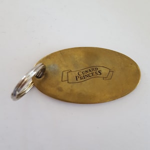 May include: A gold-colored oval keychain with a silver key ring. The keychain has the text "CUNARD PRINCESS" engraved on it.