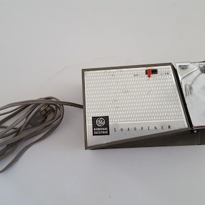 May include: A vintage General Electric electric knife sharpener with a silver and gray body. The sharpener has a power cord and a switch with an on and off setting. The text "SHARPENER" is printed on the front of the device.