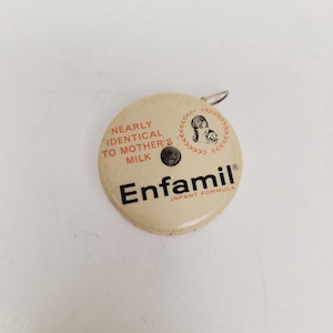 May include: A round, off-white Enfamil infant formula button with a silver metal loop. The button features the text "NEARLY IDENTICAL TO MOTHER'S MILK" and "Enfamil INFANT FORMULA". A graphic of a mother and child is also present.