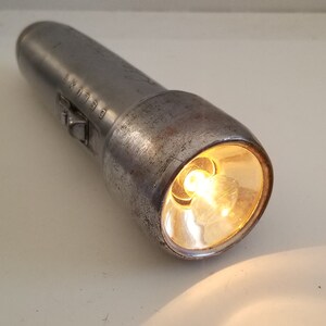 May include: A silver metal flashlight with a clear glass lens and a light bulb shining inside. The flashlight has the word "DELUXE" engraved on the side.