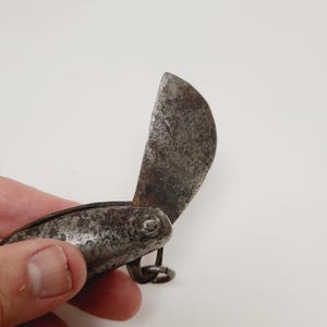 May include: An antique, small folding knife with a curved blade. The blade is open, revealing a dark, aged metal with signs of wear. The handle is also metal, with a small ring at the end.