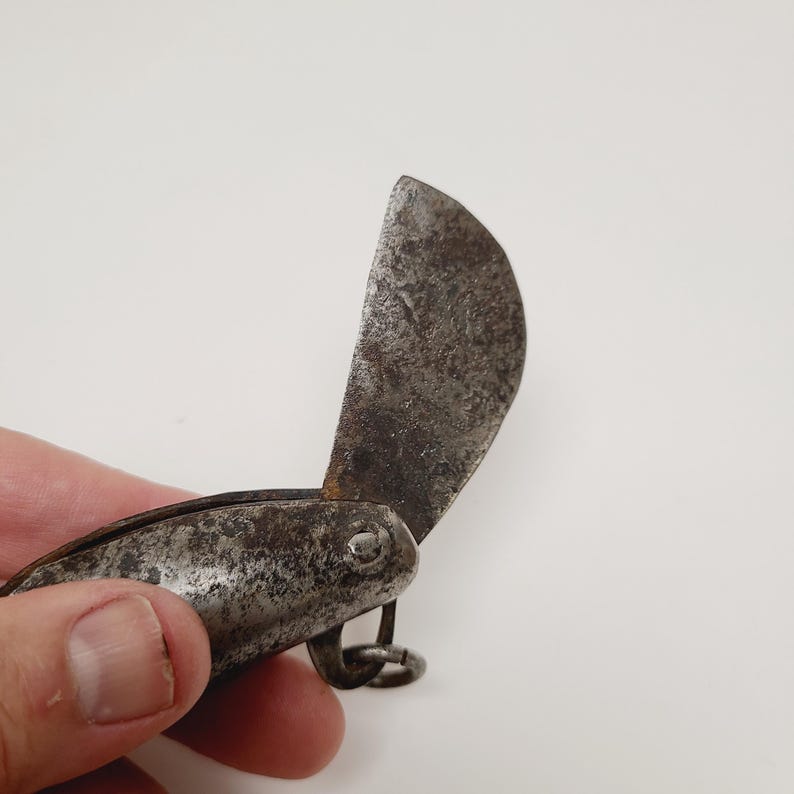 May include: An antique, small folding knife with a curved blade. The blade is open, revealing a dark, aged metal with signs of wear. The handle is also metal, with a small ring at the end.