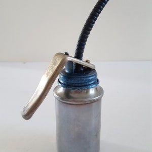 Puede incluir: Una lata de aceite plateada con una tapa azul y un largo pico flexible negro y plateado. La lata tiene una palanca plateada en el lateral.