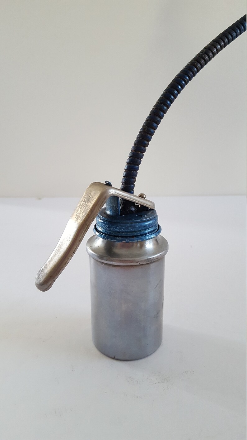 Puede incluir: Una lata de aceite plateada con una tapa azul y un largo pico flexible negro y plateado. La lata tiene una palanca plateada en el lateral.