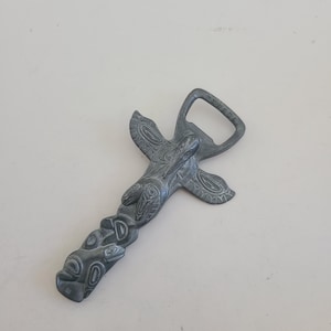 May include: A silver bottle opener shaped like a totem pole with a bird design. The opener has a traditional design with a bird on top and a series of carved figures below.