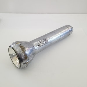 May include: A vintage silver flashlight with a ribbed head and a cylindrical body. The flashlight has a switch and the text "Lightmaster Searchlight" is visible on the side. The metal shows signs of wear.