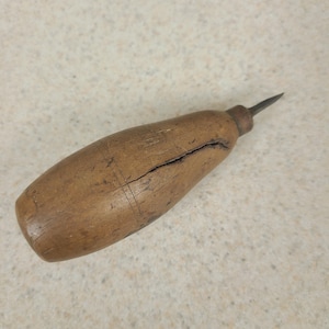 May include: A hand tool featuring a thick, barrel-shaped wooden handle with a prominent longitudinal crack and a dark, weathered finish. A short, pointed metal awl tip extends from a metal ferrule at the narrow end of the wooden handle.