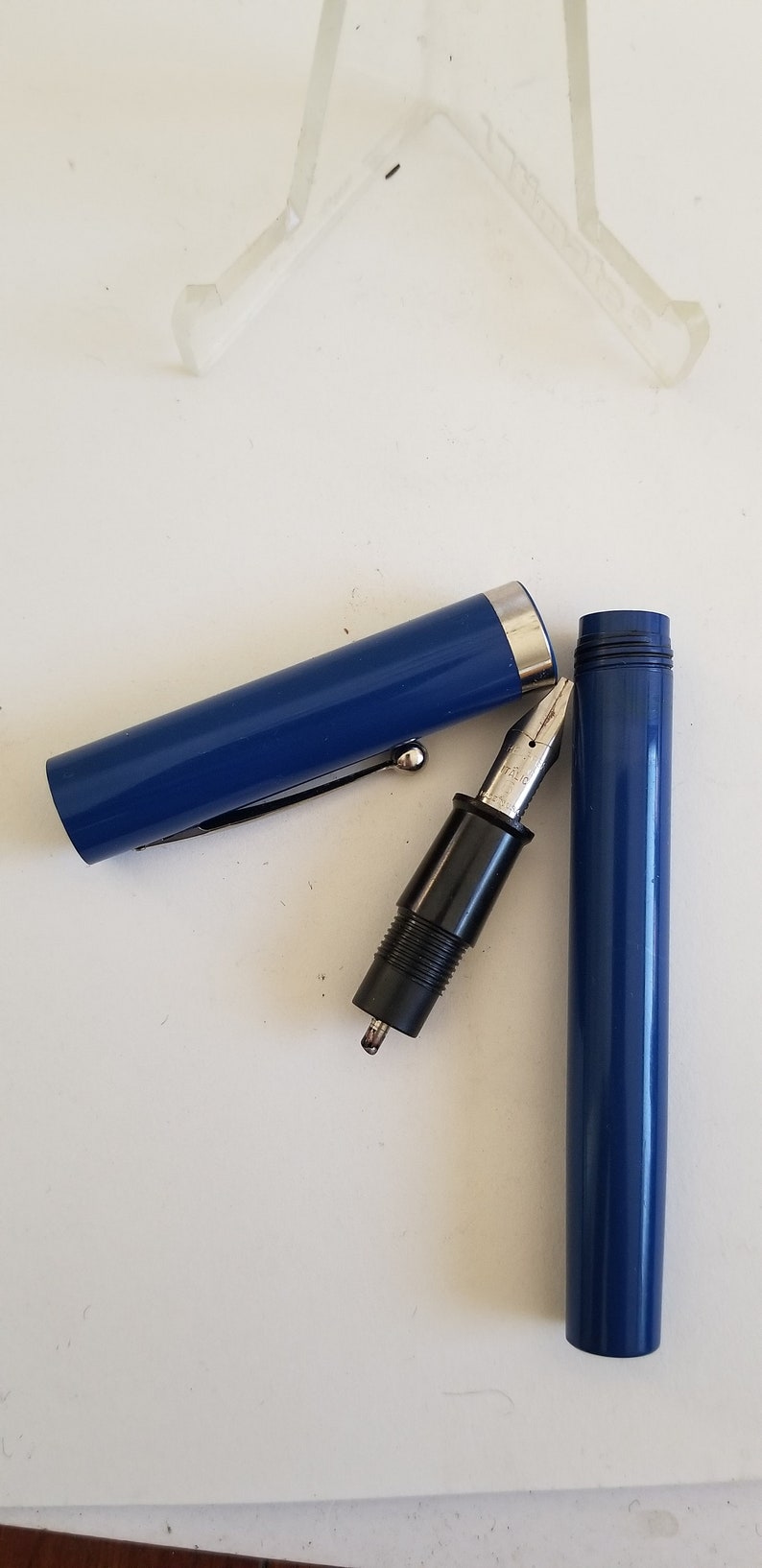 May include: A disassembled blue fountain pen with a silver nib and black grip. The pen's cap and barrel are separated, revealing the pen's internal components. A silver clip is attached to the cap.