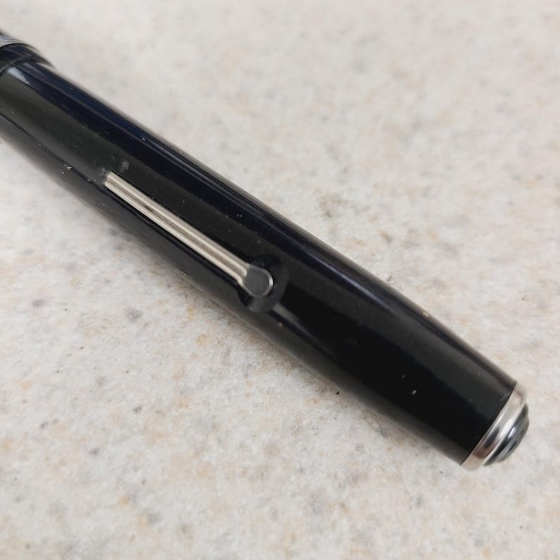 May include: Close-up of a black pen with a silver clip and accents. The pen has a glossy finish and a rounded end. The pen is lying on a light-colored surface.