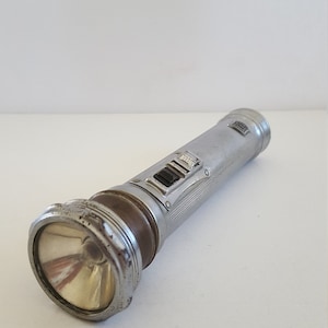 May include: A silver flashlight with a brown rubber grip and a sliding switch. The flashlight has a textured metal body and a round, clear lens.