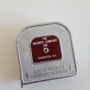 Puede incluir: Un flex&oacute;metro vintage de metal plateado con una etiqueta roja que dice "THE MAURER COMPANY INC. ROCHESTER, N.Y.". El flex&oacute;metro tiene forma curva y el texto "ADD 2 INCHES FOR INSIDE MEASURE" est&aacute; grabado en relieve.