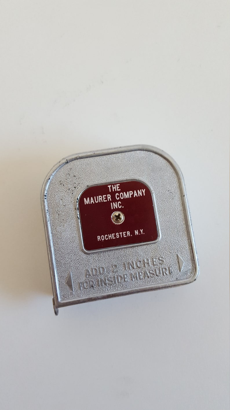 Puede incluir: Un flex&oacute;metro vintage de metal plateado con una etiqueta roja que dice "THE MAURER COMPANY INC. ROCHESTER, N.Y.". El flex&oacute;metro tiene forma curva y el texto "ADD 2 INCHES FOR INSIDE MEASURE" est&aacute; grabado en relieve.