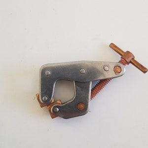 May include: A silver metal clamp with a copper-colored handle and a threaded screw. The clamp has two square copper-colored pads.