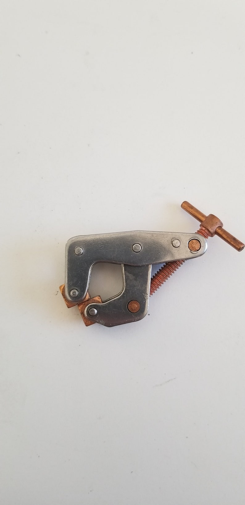 May include: A silver metal clamp with a copper-colored handle and a threaded screw. The clamp has two square copper-colored pads.