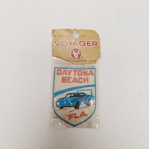 May include: A rectangular embroidered patch in a clear plastic sleeve with a cardboard header labeled A PRODUCT OF VOYAGER. The patch features a blue race car with the number 43, the text DAYTONA BEACH in red, and the abbreviation FLA. in red.