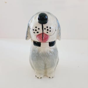 May include: A ceramic dog cookie jar with a gray and white speckled glaze. The dog has black eyes, a black nose, and a pink tongue sticking out. The dog is wearing a black collar with a white stripe.