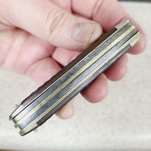 以下が含まれることがあります： 濃い茶色のハンドルと複数の刃が付いたヴィンテージのポケットナイフ。ナイフは真鍮と銀色の金属アクセントが特徴です。ハンドルは摩耗しているように見え、年齢と使用を示唆しています。ナイフは閉じられ、手に持たれています。