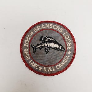 May include: Round patch with a gray center featuring a black and white fish illustration. The patch has a red border and text that reads "BRANSONS LODGE GREAT BEAR LAKE N.W.T. CANADA".