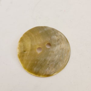 May include: A large, round button with two holes. The button has a pearlescent, olive-green color with a slightly textured surface. The button is likely made of natural material, such as shell or mother-of-pearl, and is suitable for sewing projects.