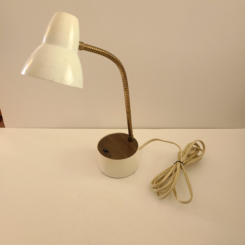 May include: A vintage white desk lamp with a flexible brass neck and a round base. The lamp has a white lampshade and a cord with a plug. The base has a wooden top.