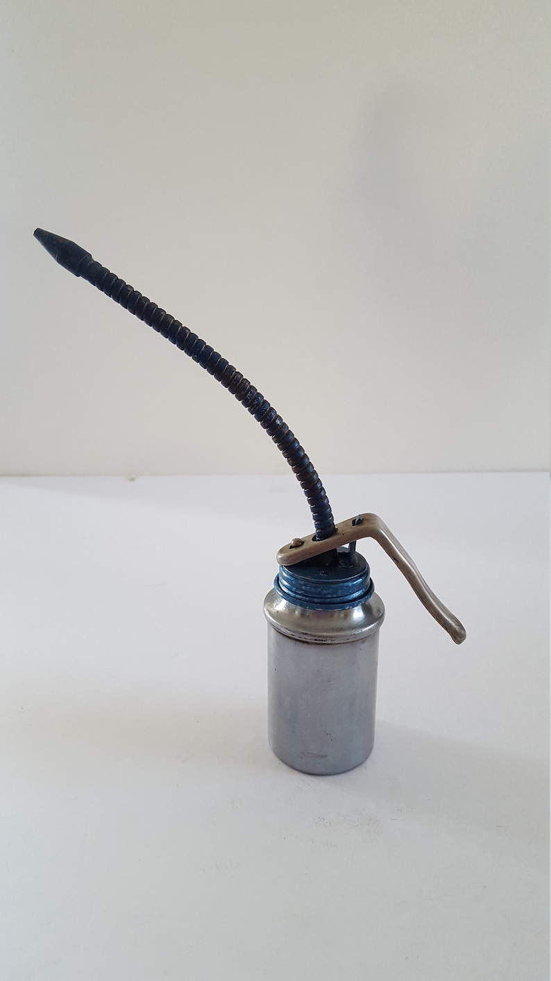 Puede incluir: Una lata de aceite plateada con un pico flexible negro y una manija de palanca. La lata tiene una tapa azul.