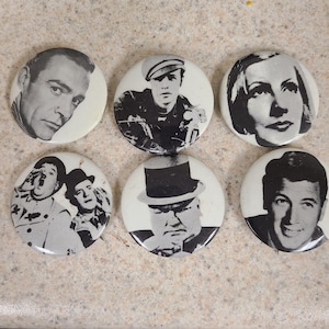 May include: Six round, white pinback buttons with black and white portraits of famous actors and personalities. The buttons feature images of Sean Connery, Marlon Brando, Greta Garbo, Laurel and Hardy, Winston Churchill, and Rock Hudson.