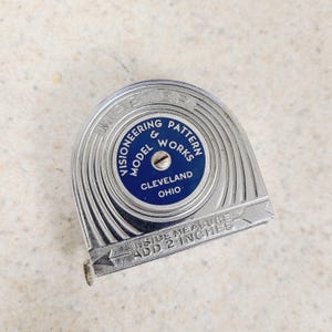 May include: A silver-toned metal tape measure with a circular blue center featuring the text MASTER, VISIONEERING PATTERN & MODEL WORKS, CLEVELAND OHIO. The casing includes the embossed instruction INSIDE MEASURE ADD 2 INCHES.