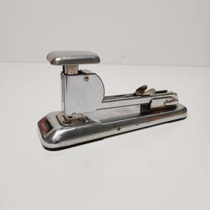 May include: A vintage chrome stapler with a rectangular base and a curved top. The stapler has a lever and a flat top for applying pressure. The metal has a shiny, reflective surface, and the stapler is set against a white background.