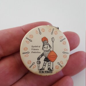 May include: A white and orange round measuring tape with a cartoon knight holding a sword and shield. The text on the tape reads "Symbol of Vitamin Protection" and "Sir Vi-Sol".