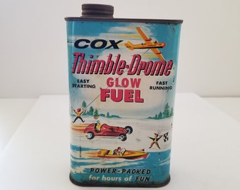Peut inclure: Une boîte en métal vintage de carburant pour modèles réduits Cox Thimble-Drome Glow Fuel. La boîte est bleue avec un avion jaune et des voitures de course rouges et blanches. Le texte sur la boîte indique "Cox Thimble-Drome Glow Fuel" et "Power-Packed for hours of fun".