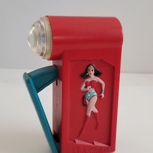 May include: A red plastic flashlight with a Wonder Woman character image on the side. The flashlight has a blue handle and the text "FLASHMITE" is printed on the bottom.