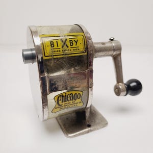May include: A vintage metal pencil sharpener with a hand crank and a black knob. It features a clear plastic casing with yellow labels reading BIXBY GRAND RAPIDS, MICH. and Chicago MADE BY AUTOMATIC PENCIL SHARPENER CO. CHICAGO, U.S.A. on the front.