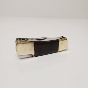 May include: A closed pocket knife with a dark wood handle and brass-colored metal ends. The blade is silver-colored. The knife is lying horizontally on a white surface.