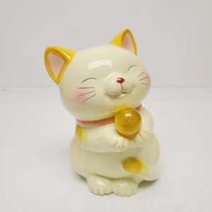 May include: A ceramic Maneki Neko figurine in a seated pose. The cat is primarily cream-colored with yellow accents on the ears and tail. It holds a golden orb and wears a pink collar. The cat has a smiling expression with rosy cheeks.
