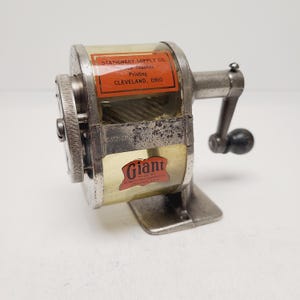 May include: Vintage silver-toned pencil sharpener with a clear plastic window and a black handle. The sharpener has a crank and a base. The labels read "STATIONERY SUPPLY CO." and "Giant".