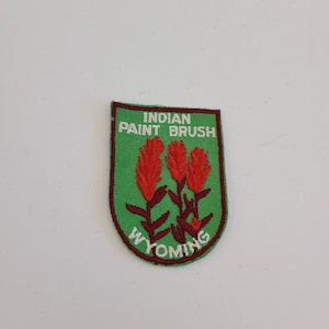 May include: A green and maroon embroidered patch featuring the words "Indian Paint Brush" and "Wyoming". The patch has a green background with three red flower illustrations.