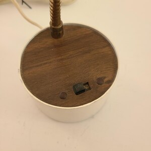 May include: A vintage desk lamp with a white base and a wooden top. The lamp features a flexible, brass-colored gooseneck and a small on/off switch. The base is round and has a retro aesthetic.