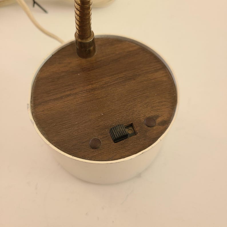 May include: A vintage desk lamp with a white base and a wooden top. The lamp features a flexible, brass-colored gooseneck and a small on/off switch. The base is round and has a retro aesthetic.