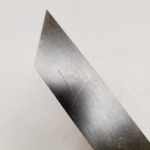 May include: A silver-colored metal utility knife blade with an angled tip. The surface features an etched Stanley logo inside a heart shape, with the text MADE IN U.S.A. positioned directly below the logo. The metal shows light surface scratches.