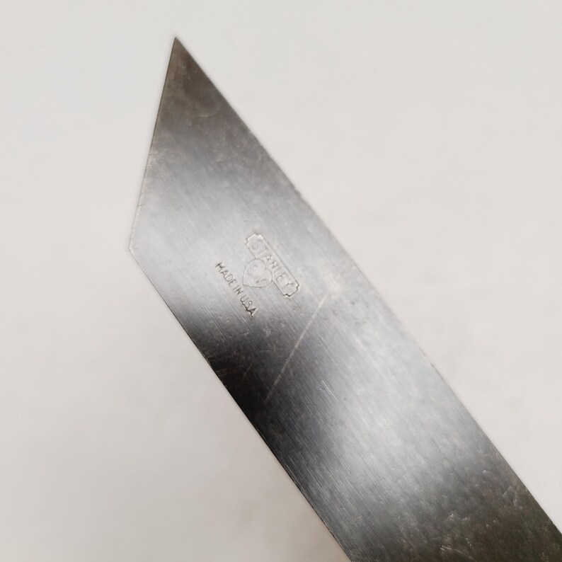 May include: A silver-colored metal utility knife blade with an angled tip. The surface features an etched Stanley logo inside a heart shape, with the text MADE IN U.S.A. positioned directly below the logo. The metal shows light surface scratches.