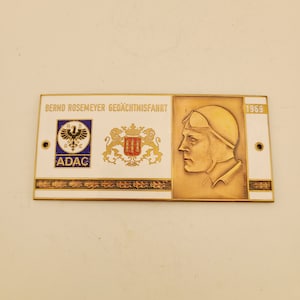 May include: A white and gold metal plaque with the text "BERND ROSEMEYER GEDÄCHTNISFAHRT 1969" and a black and white logo for ADAC. The plaque also features a coat of arms with a lion and a crest.