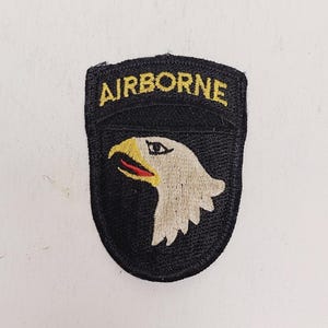 May include: A black patch with the word "AIRBORNE" in yellow lettering at the top. Below the text is a white eagle head with a yellow beak. The patch is a military insignia.
