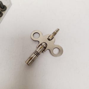 May include: A silver-colored clock key with a circular handle on each end and a winding mechanism in the center. The key is designed to wind a mechanical clock and is made of metal.
