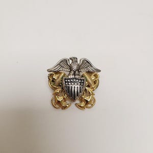 May include: A silver and gold metal pin featuring a detailed eagle with outstretched wings atop a shield with vertical stripes. The shield is flanked by gold anchors and laurel wreaths, creating a military or nautical theme.
