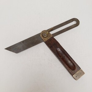 May include: A sliding T-bevel tool featuring a dark brown wooden handle with a recessed grip, a brass locking nut and end cap, and a flat steel blade with a slotted end. The metal components show a weathered patina from age and use.