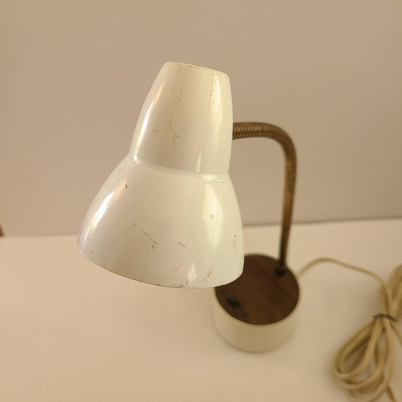 May include: A vintage white desk lamp with a brass-colored flexible neck and a round wooden base. The lamp has a classic, retro design, with a white lampshade and a cord for power.